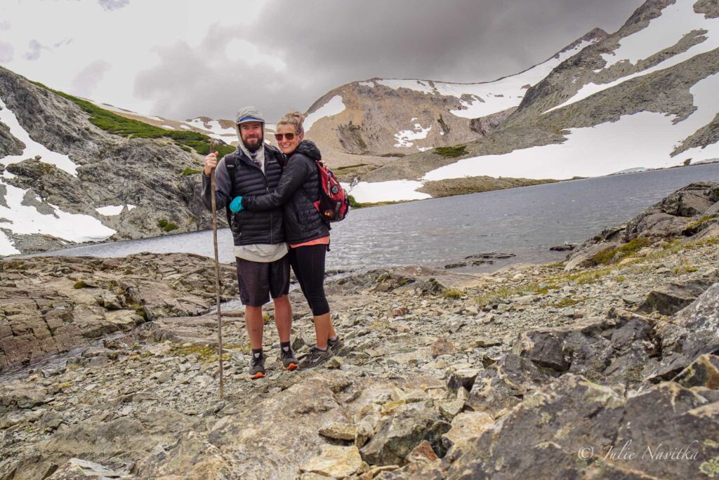 Image of a couple together in front of a mountain lake with rocky banks and small amount of snow.