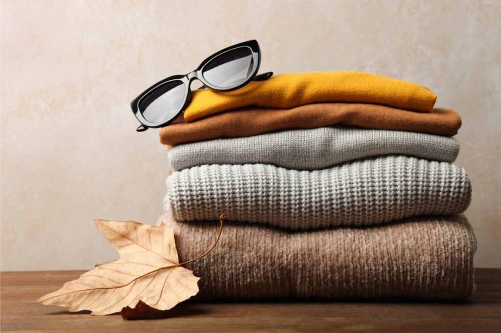 Image of four neutral coloured sweaters and tops folded in a neat pile with a pair of sunglasses on top and a leaf resting against them. What to pack for a year of travel includes sustainable clothing.