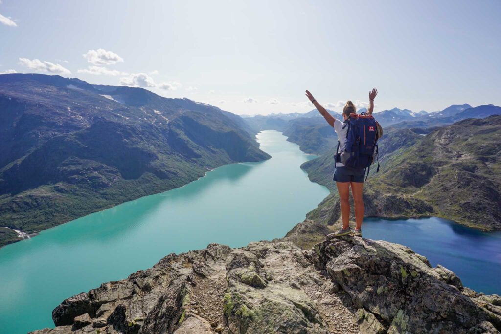Image of a hiker with their arms in the ari wearing a large backpack standing on a rocky ledge high above a view of a turquoise lake in Norway.