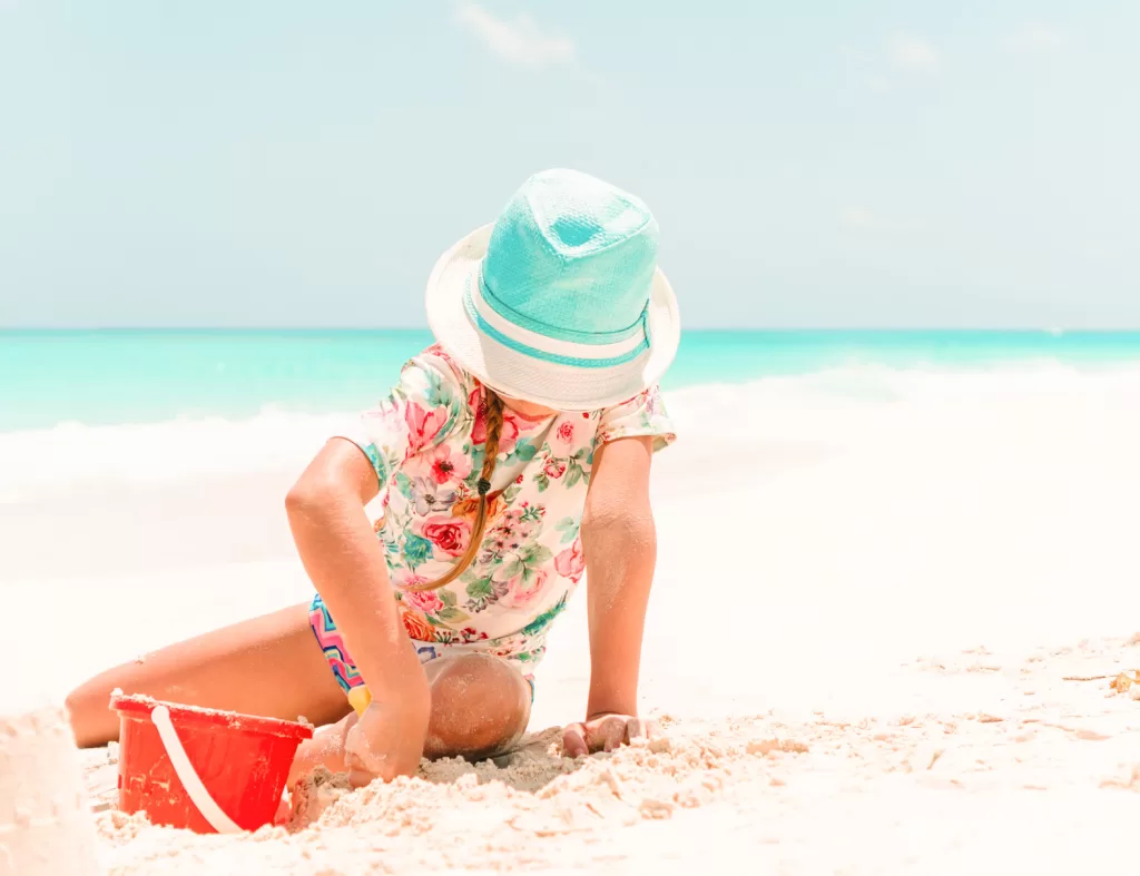Image of a young girl wearing a bucket hat playing with a shovel and pail on the beach. Recreating sustainably at the beach is important too!