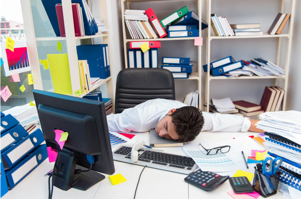 Image of a man with his head down on his desk, exhausted amidst piles of clutter in his office.