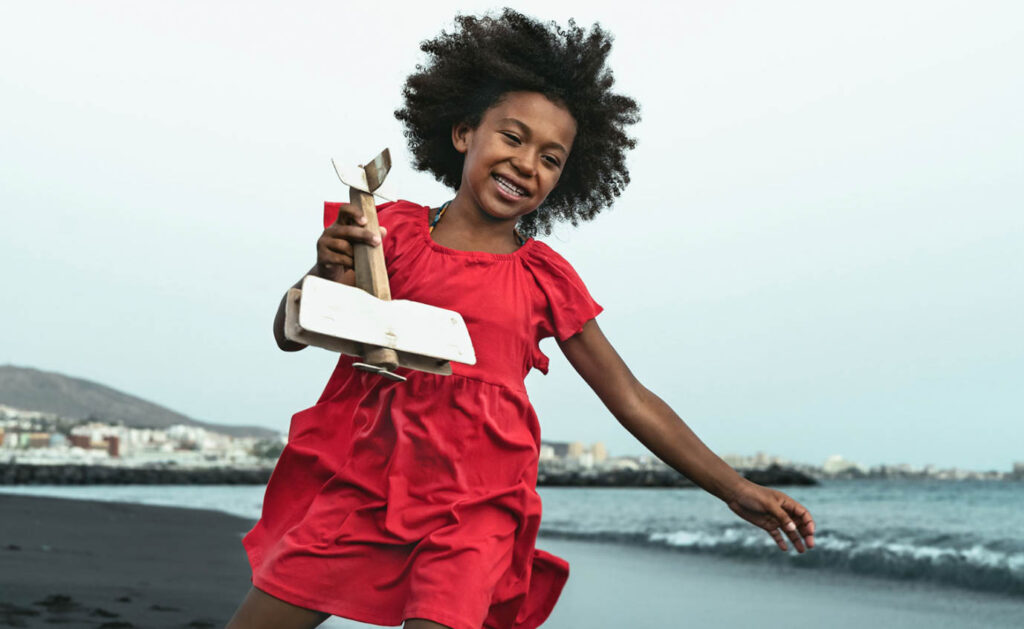 Image of a young girl wearing a red dress running on the beach with a wooden plane in her hand.