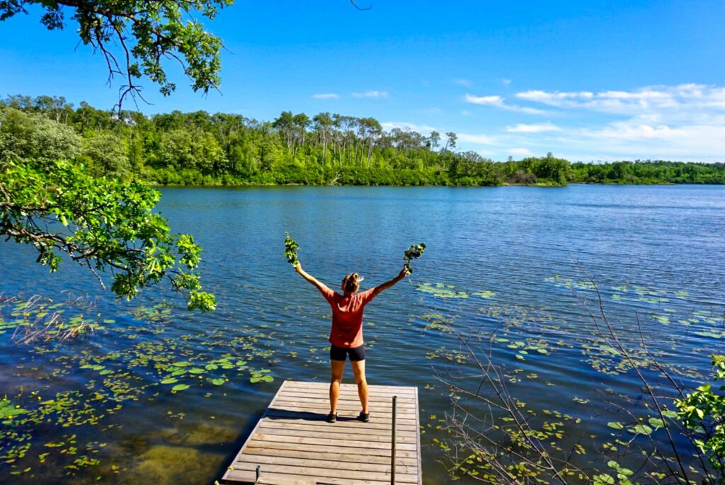 Image of a hiker standing on a dock at a lake with their arms spread wide holding some leafy branches. You don't need fancy technical gear to build a sustainable hiking capsule wardrobe.