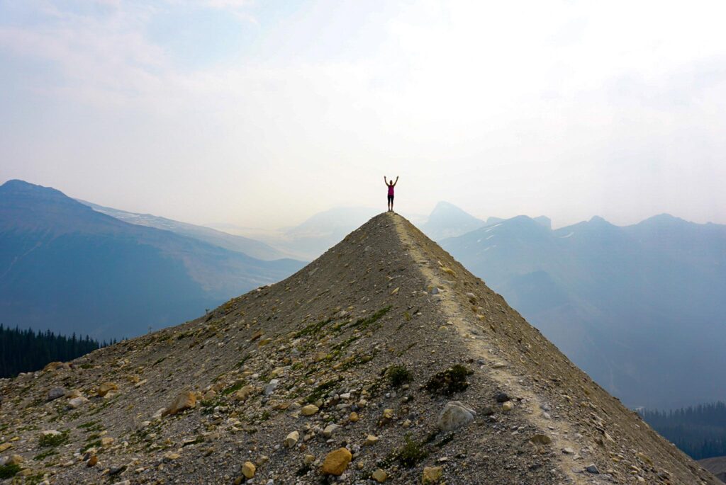 Image of the tiny silhouette of a hiker with their arms up atop a ridge with mountains in the backgournd.