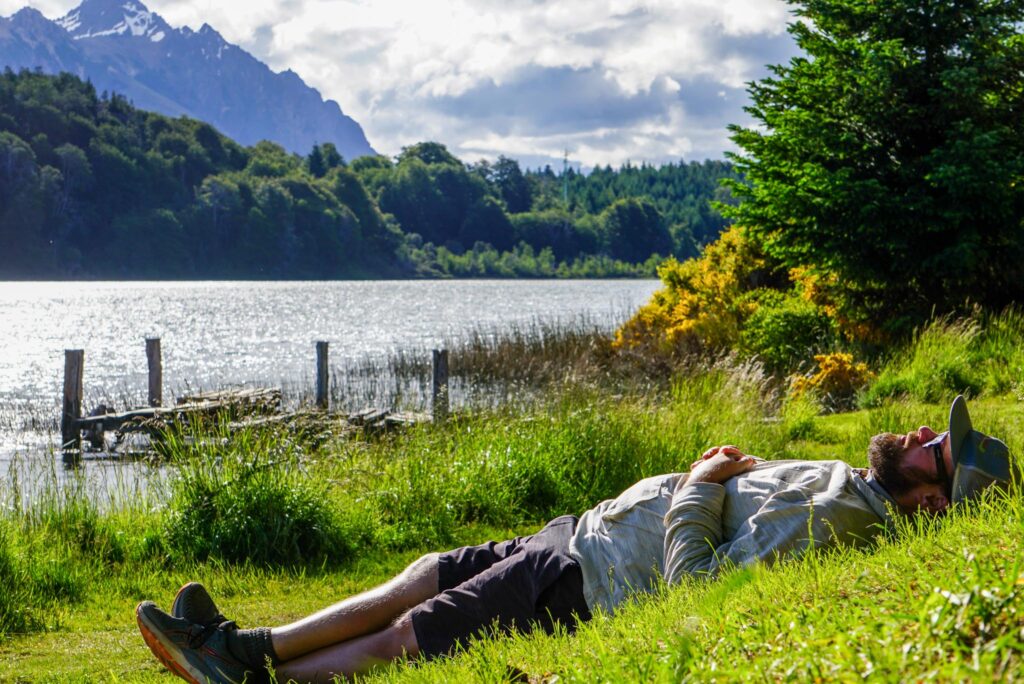 Image of a man laying in the grass by a lake in the mountains, relaxing and looking at the sky.