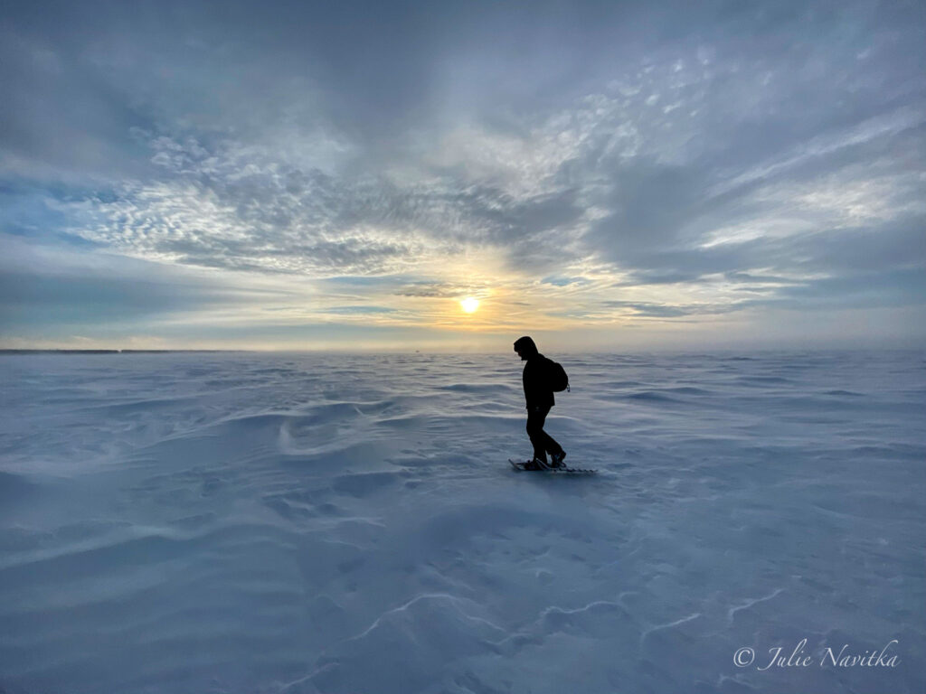 Silhouetted image of a snowshoer on a frozen lake at sunset. Some outdoor enthusiasts will need to include extreme weather gear in their hiking capsule wardrobe.