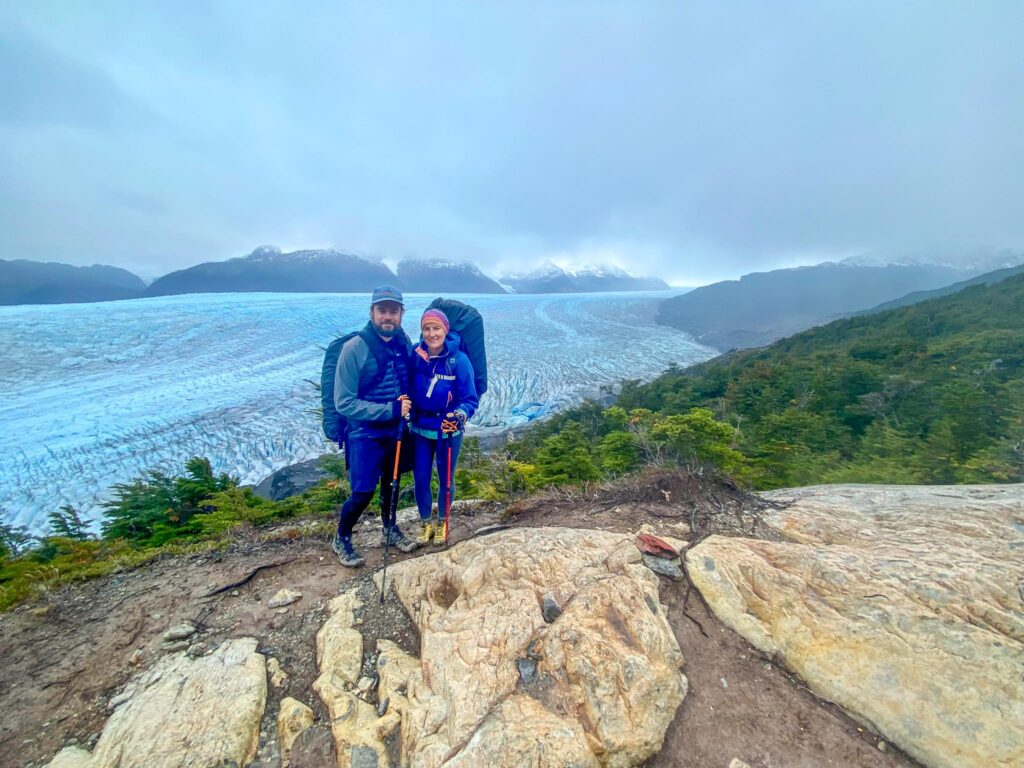 Image of a hiking couple wearing warmer hiking clothing taken in front of a massive glacier in Chile. If you hike in colder climates, your hiking capsule wardrobe will require layering items.