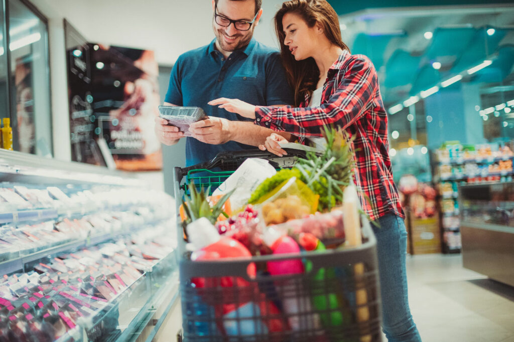 Image of a couple with a shopping cart filled with produce looking at a package at the deli counter. Buying in-season produce and meat alternatives can help you shop sustainably on a budget.