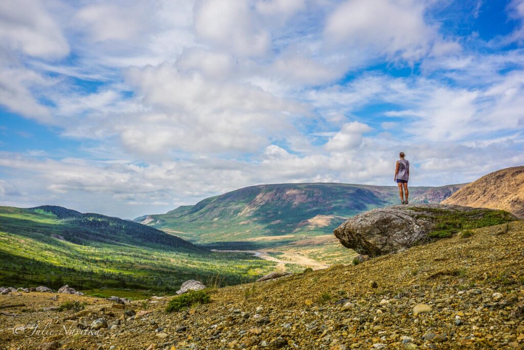 Image of a hiker atop a boulder looking out over a vast landscape of rolling hills and rocks under a blue sky scattered with clouds. My hiking capsule wardrobe contains second-hand items, like the tank top here. 