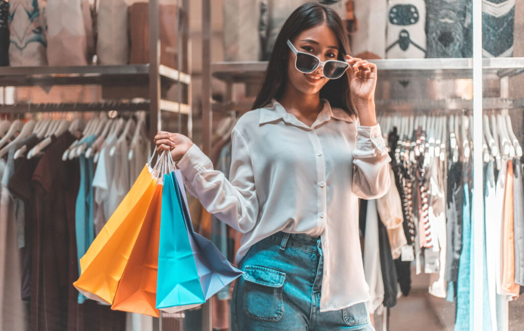 Image of a woman in a store with a few shopping bags in her hand, tipping her sunglasses to look at the camera.