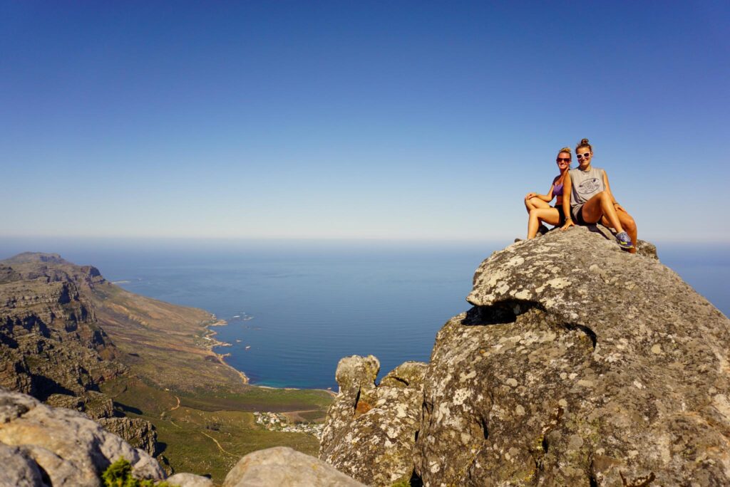 Image of two women sitting on top of a rocky outcrop in front of a view of the ocean below. Your hiking capsule wardrobe will need to include clothing for hot weather too.