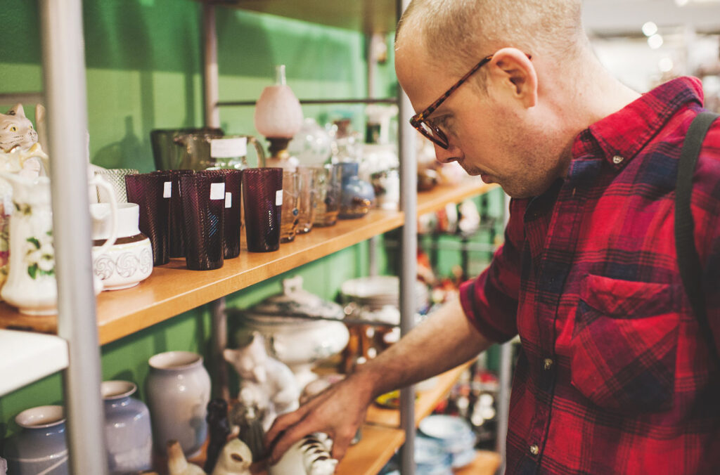 Image of a man perusing some shelves of secondhand items at a thrift store. Buying used items can help you shop sustainably on a budget.