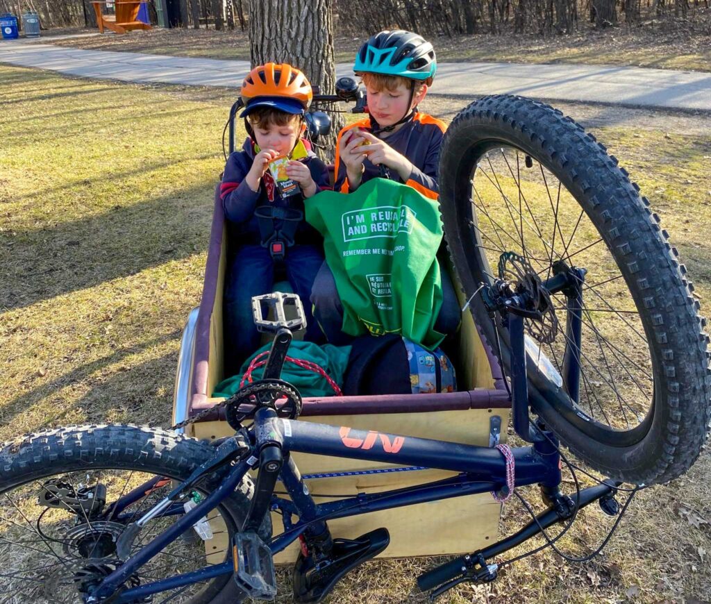 Image of two young children sitting in a bike carrier stopped to enjoy a snack in the park. Make your school drop off part of your environmentally friendly work commute.