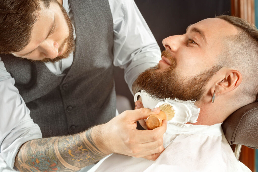 Image of a man having his beard shaved at a barber shop - the barber is applying a thick shaving soap lather to his face.