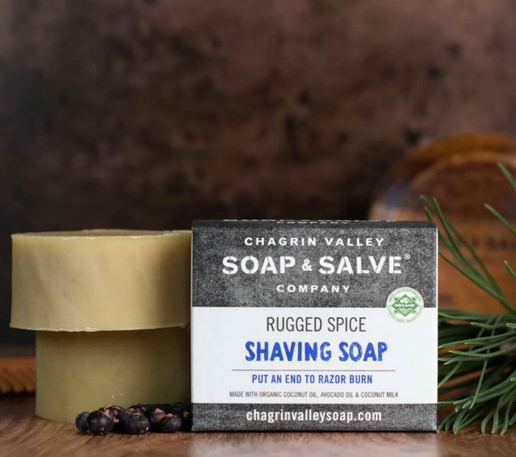 Image of one packaged shaving soap by Chagrin Valley next to two naked soaps on a wooden surface. 