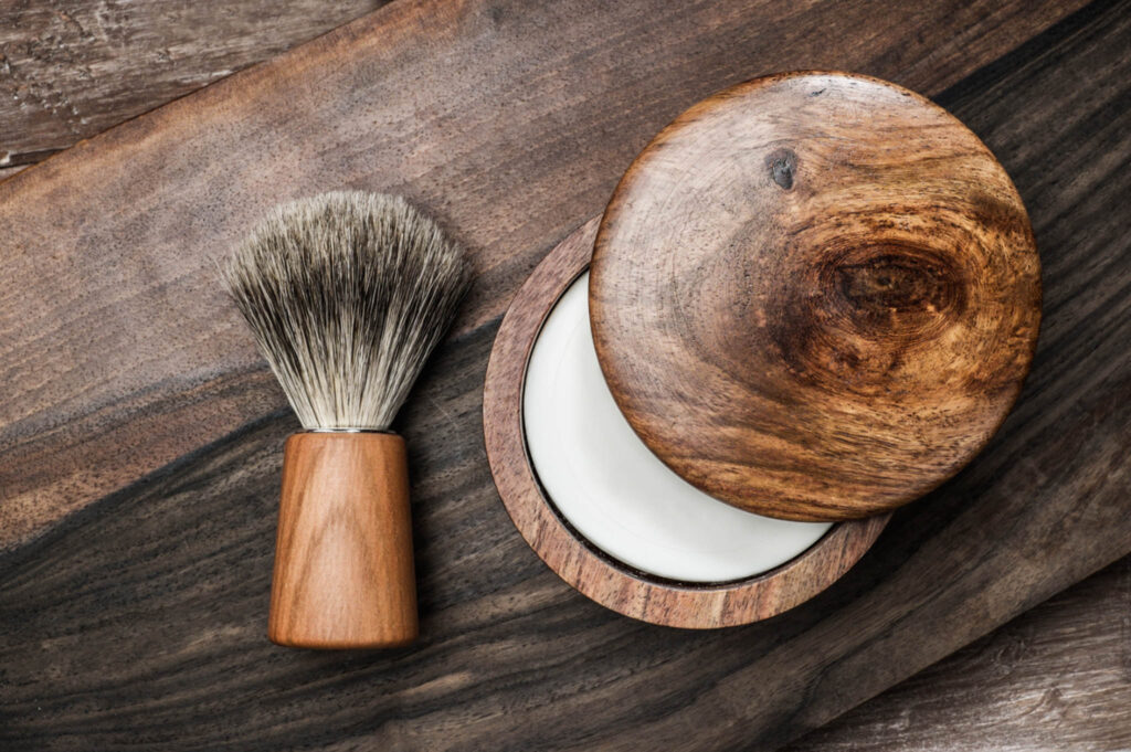 Image of a shaving soap in a wooden bowl next to a shaving brush on a wooden countertop.