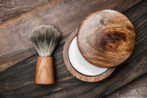 Image of a shaving soap in a wooden bowl next to a shaving brush on a wooden countertop.
