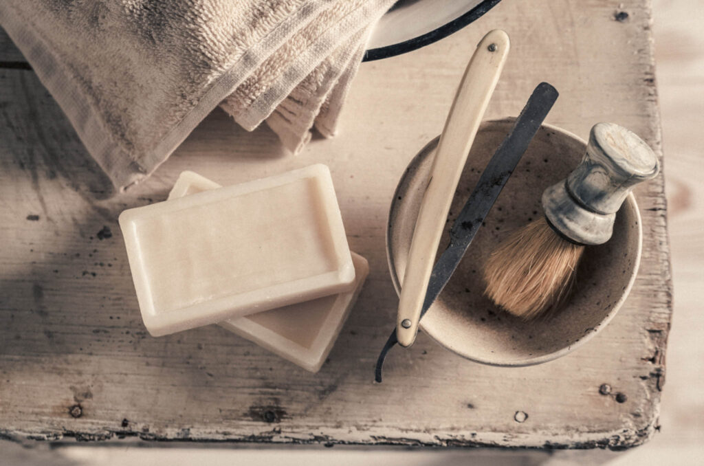 Image taken from about of two bars of soap, a shaving brush and bowl set with straight razor and towel sitting on a counter. Shaving soaps can be a great sustainable alternative to cream for sensitive skin.