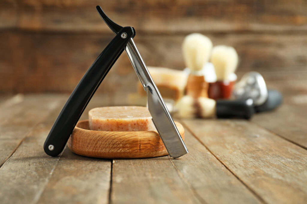 Image of a straight razor open above a shaving soap bar sitting in a wooden dish on a counter with shaving brushes in the backgournd.