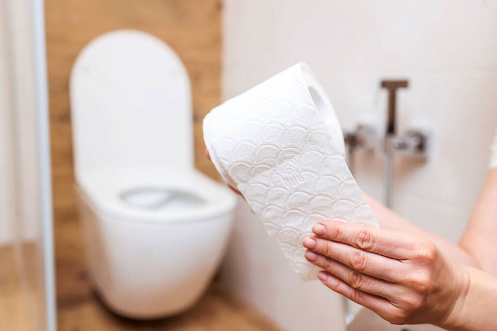 Image of a hand holding and pulling some sheets from a roll of toilet paper with a bidet in the background.
