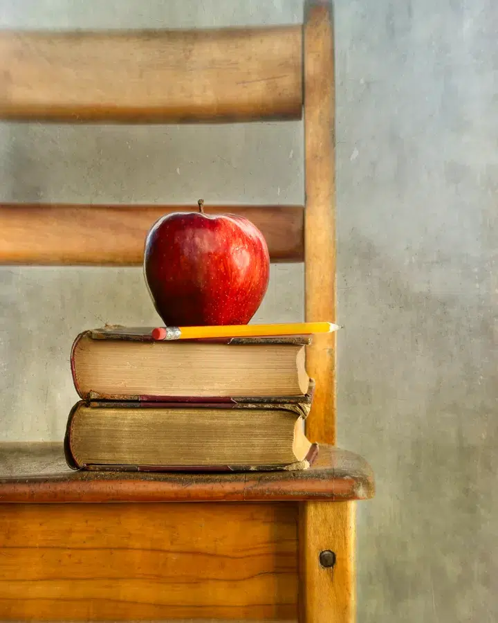 Image of a wooden chair with two books on the seat, with a red apple perched on top.