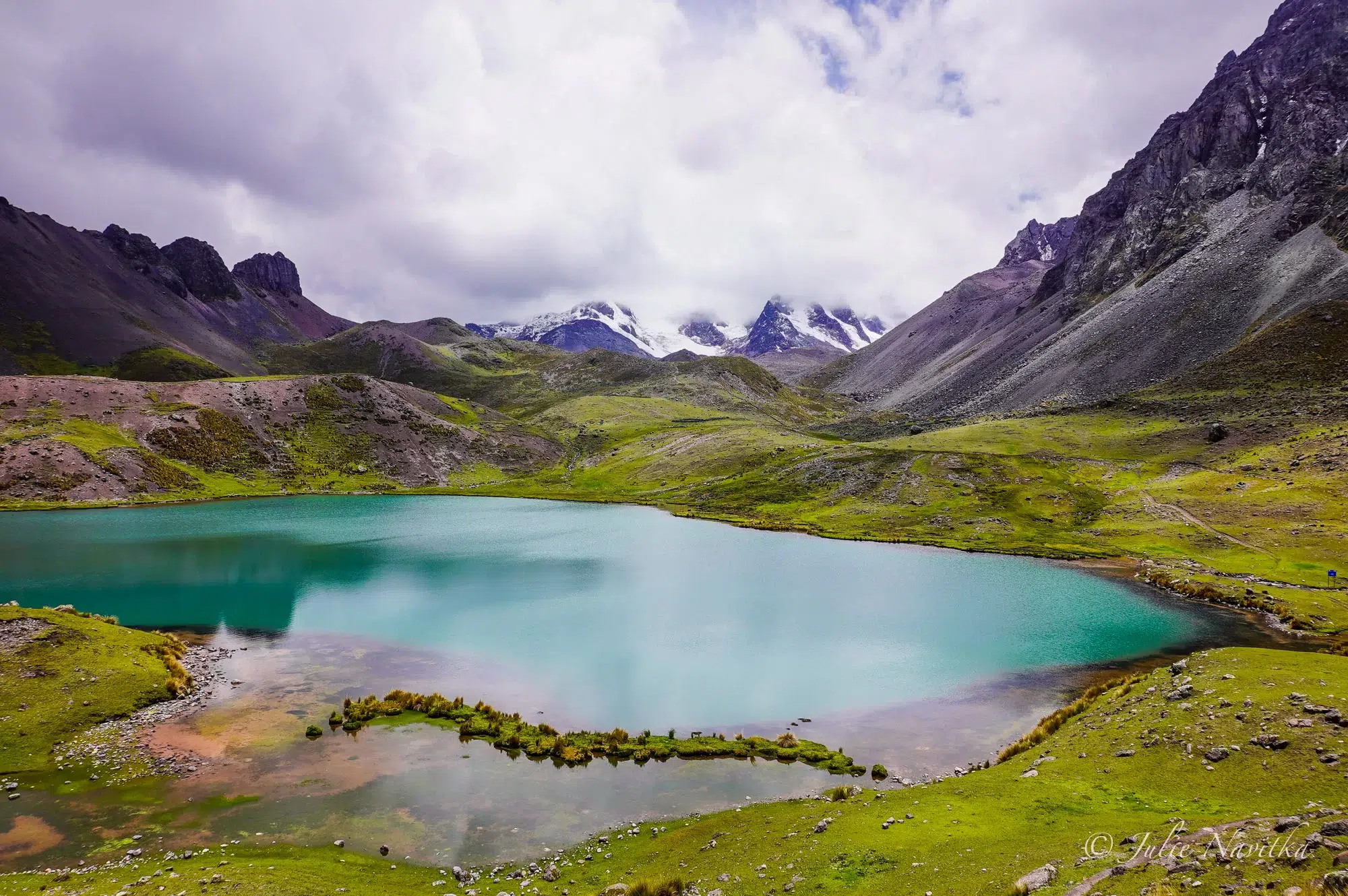 Image of a turquoise lake surrounding by lush vibrant green grasses and towering mountains near Cusco, Peru.