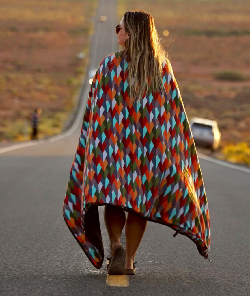 Image of a woman with the Kachula blanket from Coalatree draped over her shoulders walking down the middle of the road in what appears to be an isolated area in nature.