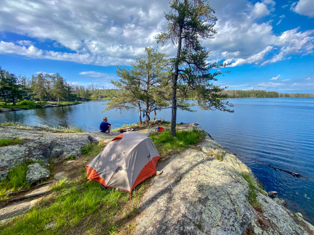 Image of a tent set up near a lake and a tree on a blue sunny day. A man site on the shoreline admiring the view.
