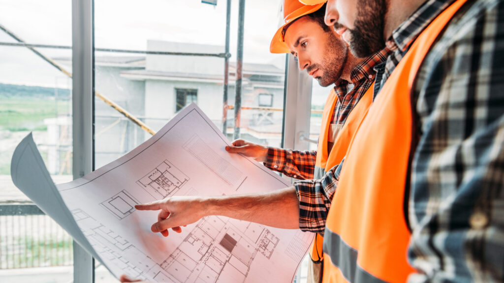 Image of two men in high visibility vests and hard hats look over blueprints together. Sustainable business operations lead to many benefits in the workplace.