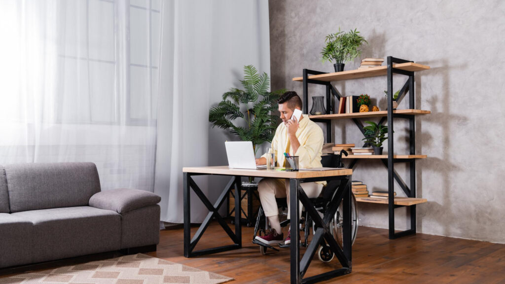 Image of a man in a wheelchair sitting at a desk in a well-lit office with a shelf containing plants behind him. Relying on natural light and renewable energy sources can lead to economic benefits for your sustainable workplace.