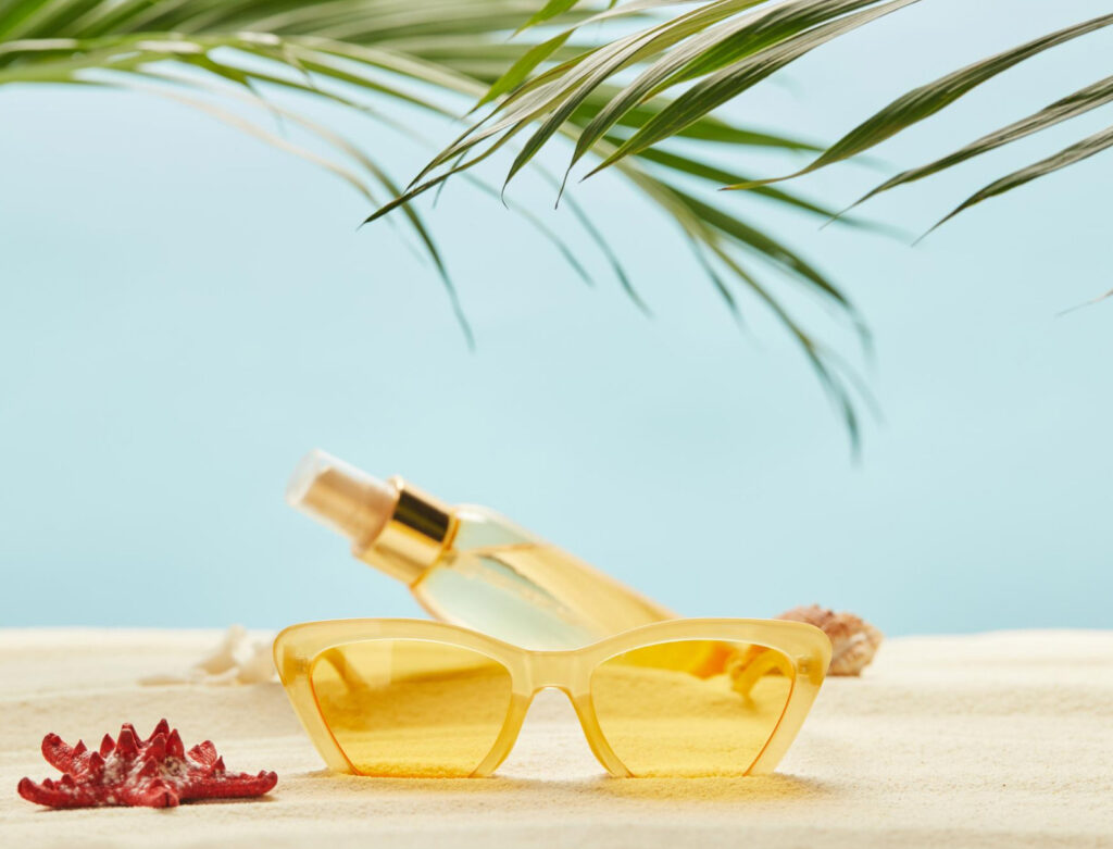 Image of a pair of sunglasses on the sand in front of a spray bottle of product. A palm frond hangs overtop with a blue sky in background.