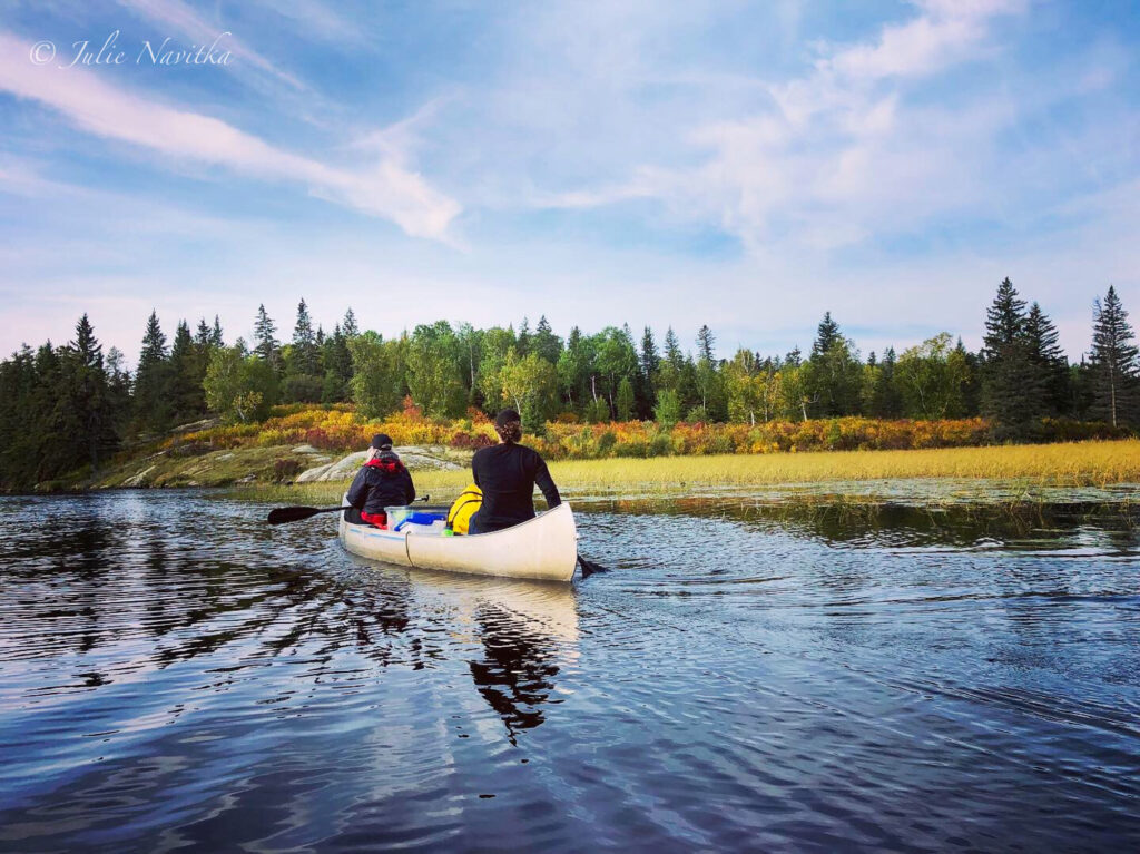Image of two people canoeing away from the photographer on a lake with a treed shoreline in the background.