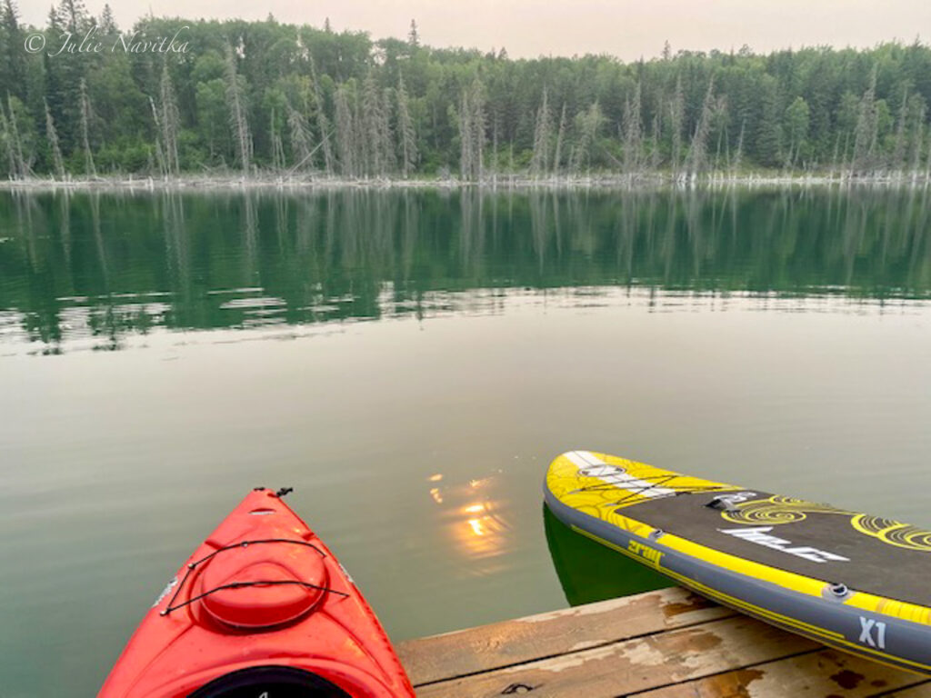 Image of a kayak and a stand-up paddleboard sitting on a dock at a lake with trees reflected in the water. Sustainable water board sports are human-powered activities!