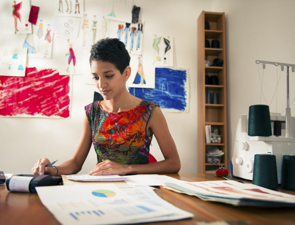 Image of a woman sitting at a desk with fashion designs on the all behind her. environmentally responsible companies have happier and more productive employees.