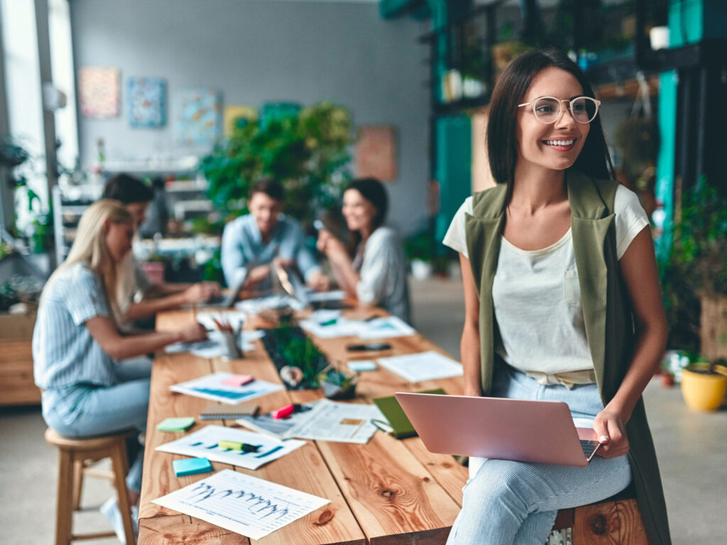 Image of a smiling woman at the end of a conference table in a sustainable workplace with coworkers seated and collaborating behind her.