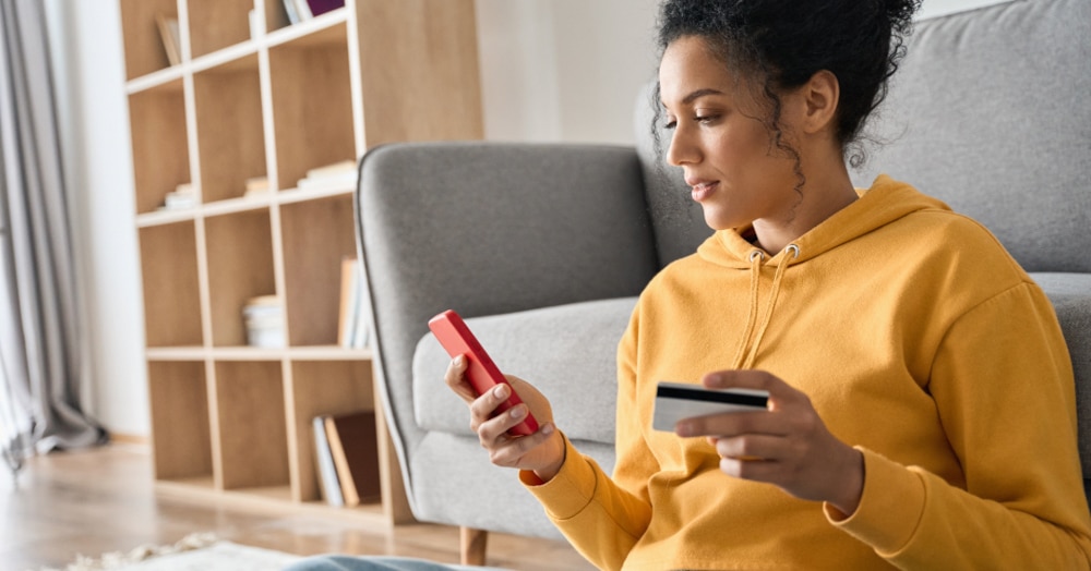 Image of a woman holding a smartphone and a credit card while she online shops in her living room.