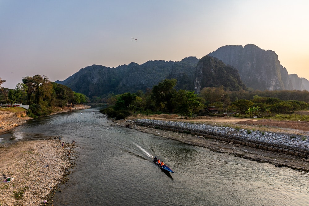 Image of a narrow boat speeding down a river with mountains in the background. Visit Vang Vieng sustainably by choosing low-impact activities. 