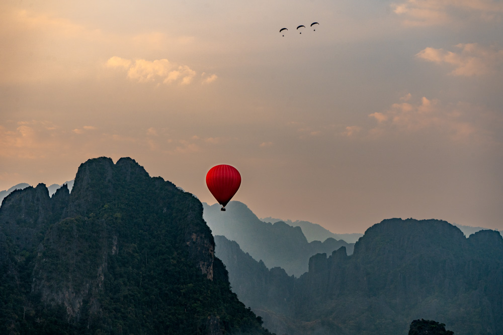 Image of a red hot air balloon floating in front of silhouetted mountains at dusk. Any Vang Vieng travel guide will include a hot air balloon ride!
