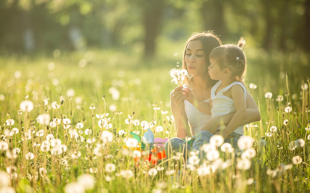 Image of a young child and her mother in a field blowing dandelion fluff.