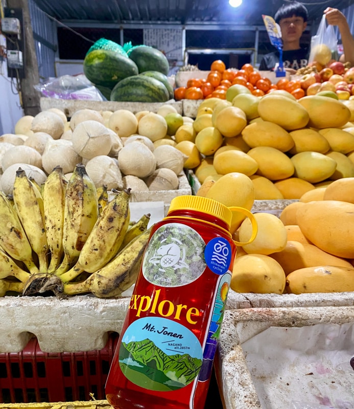 Image of a reusable water bottle in front of an array of tropical fruits at a market stall. Buying local produce can help you visit Vang Vieng sustainably and on a budget.