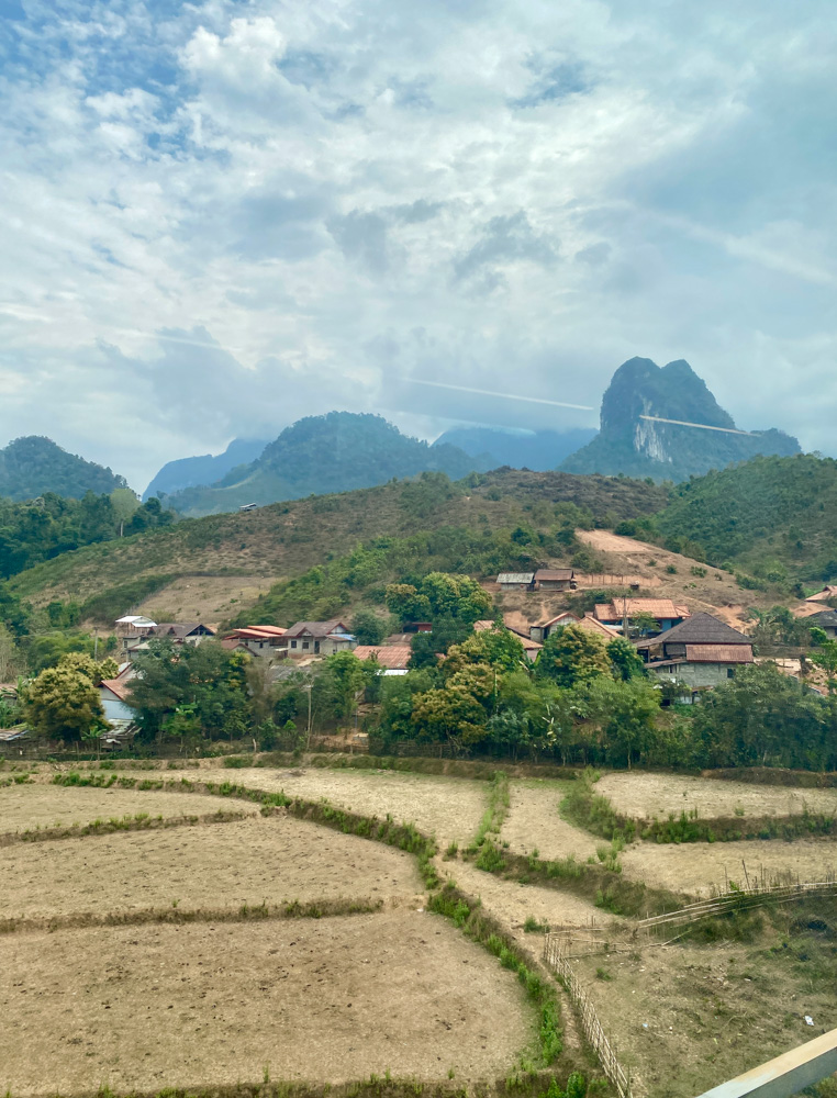 Image of rice fields, buildings, and mountains in the background in the Laos countryside.  