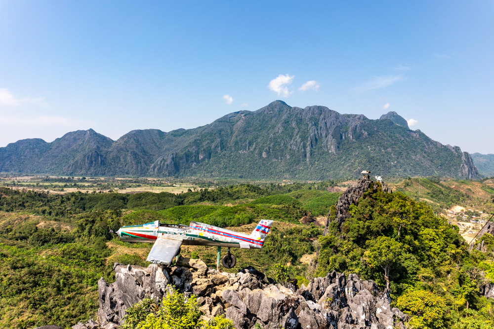Image of an old small airplane perched on top of a rocky peak with mountains in the background. 