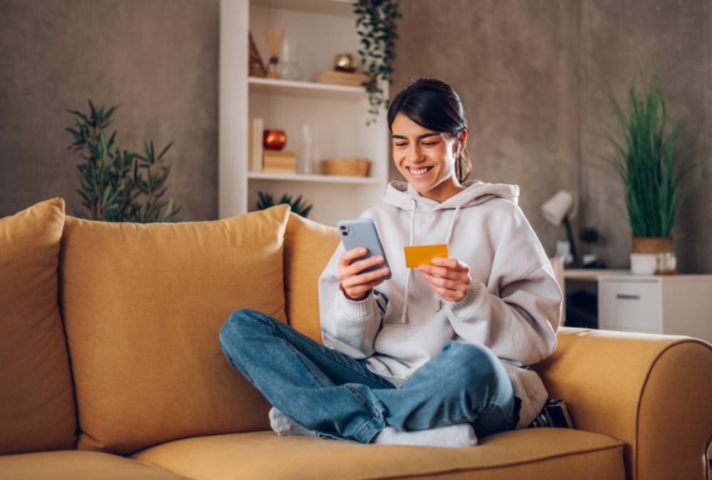 Image of a woman sitting on a couch with her smartphone and credit card in hand. 