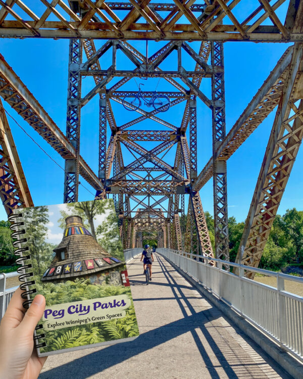 Image of the 'Peg City Parks book in front of a train bridge with cyclist crossing.