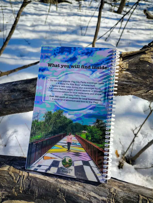 Image of the back of the 'Peg City Parks - Explore Winnipeg's Green Spaces" book on a tree, featuring scavenger hunts for 10 different Winnipeg parks.