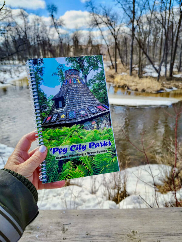 Image of the 'Peg City Parks - Explore Winnipeg's Green Spaces" book in front of the Seine River, featuring scavenger hunts for 10 different Winnipeg parks.