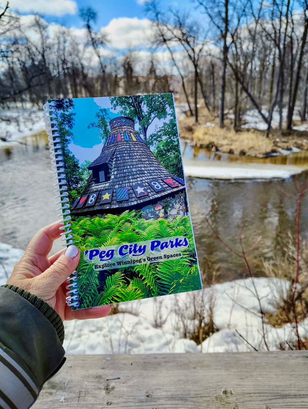 Image of the 'Peg City Parks - Explore Winnipeg's Green Spaces" book in front of the Seine River, featuring scavenger hunts for 10 different Winnipeg parks.